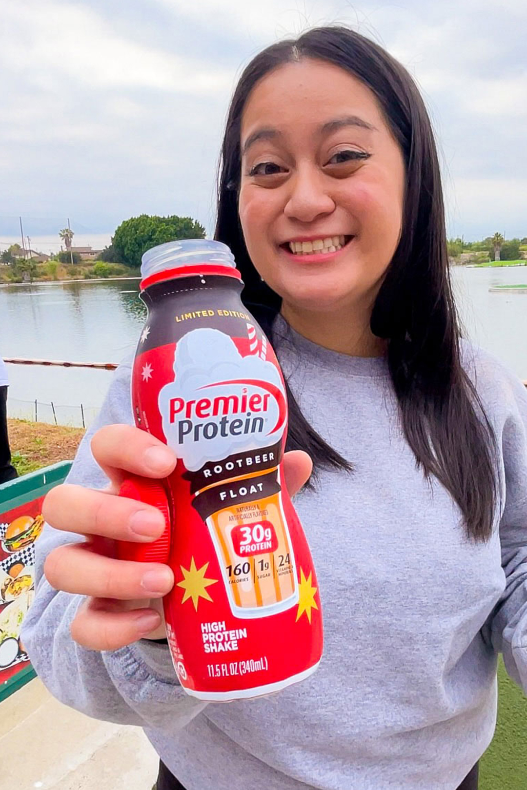 Woman holding up a bottle of Premier Protein Root Beer Float next to a body of water.