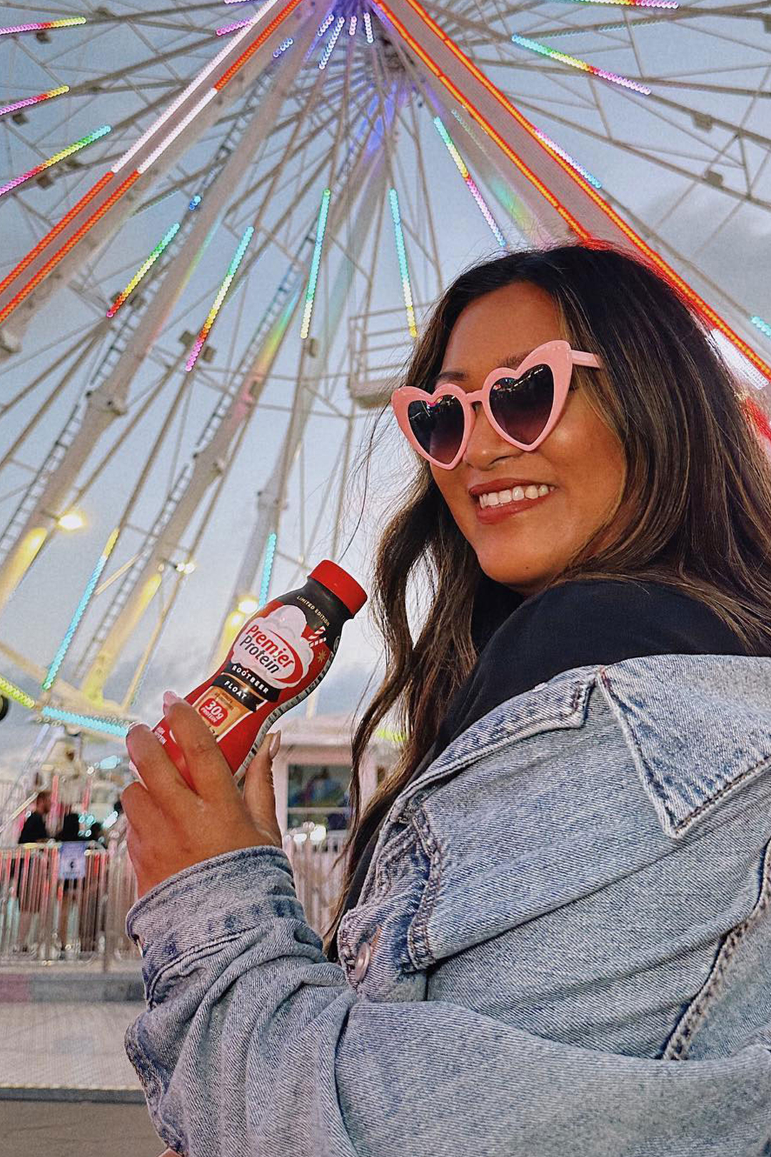 Woman in sunglasses holding a bottle of Premier Protein Root Beer Float in front of a ferris wheel.