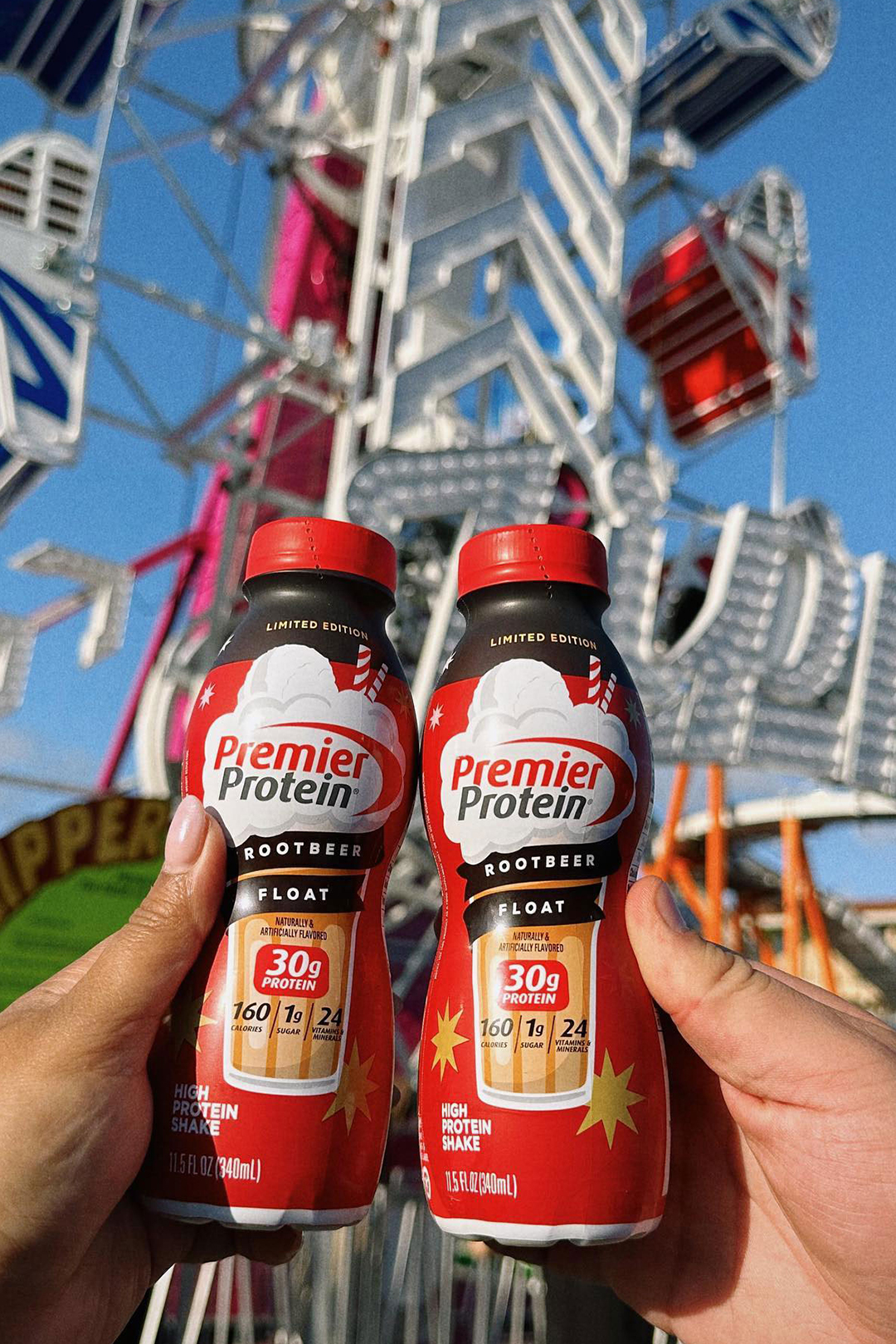 Two hands cheers-ing bottles of Premier Protein Root Beer Float shake in from of a carnival ride