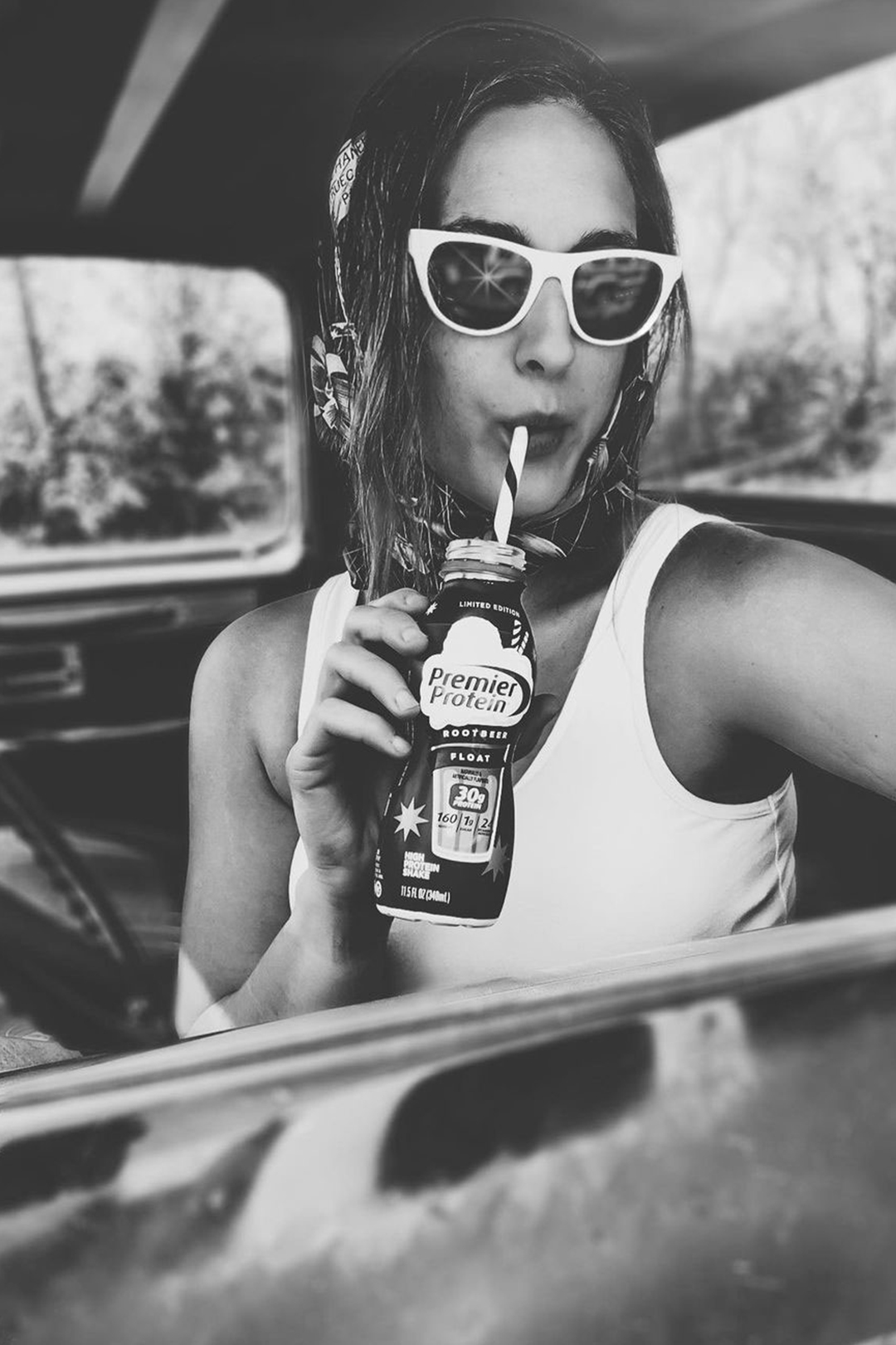 Black and white photo of a woman in sunglasses sitting in a car while drinking Premier Protein Root Beer Float shake from a straw