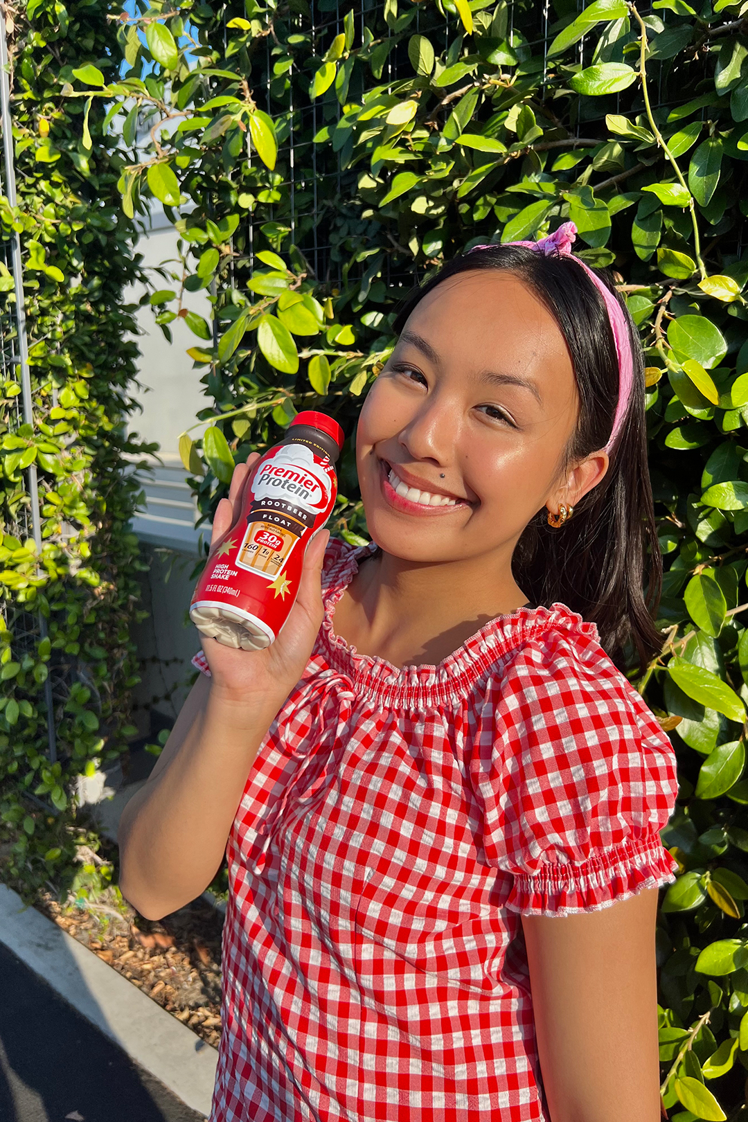 Woman smiling outdoors and holding up a bottle of Premier Protein Root Beer Float protein shake